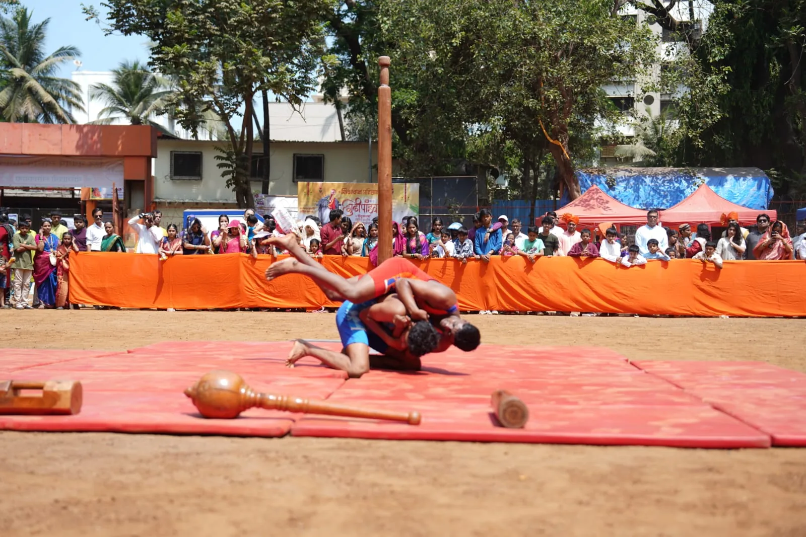 Two Kusti wrestlers grappling on the red mat, traditional gada maces resting in the foreground