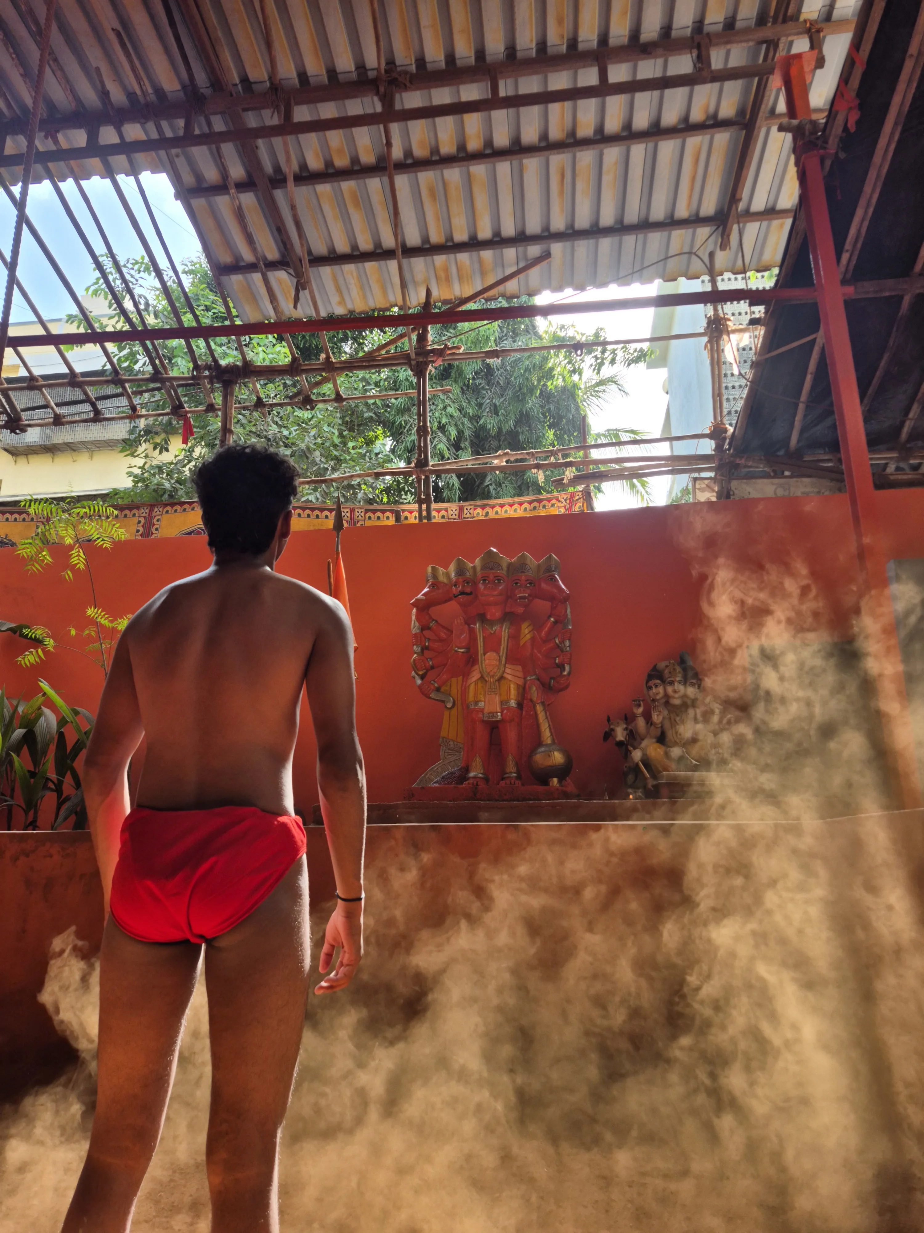 Wrestler standing in the red earth of the akhada facing the Hanuman shrine, dust hanging in the light